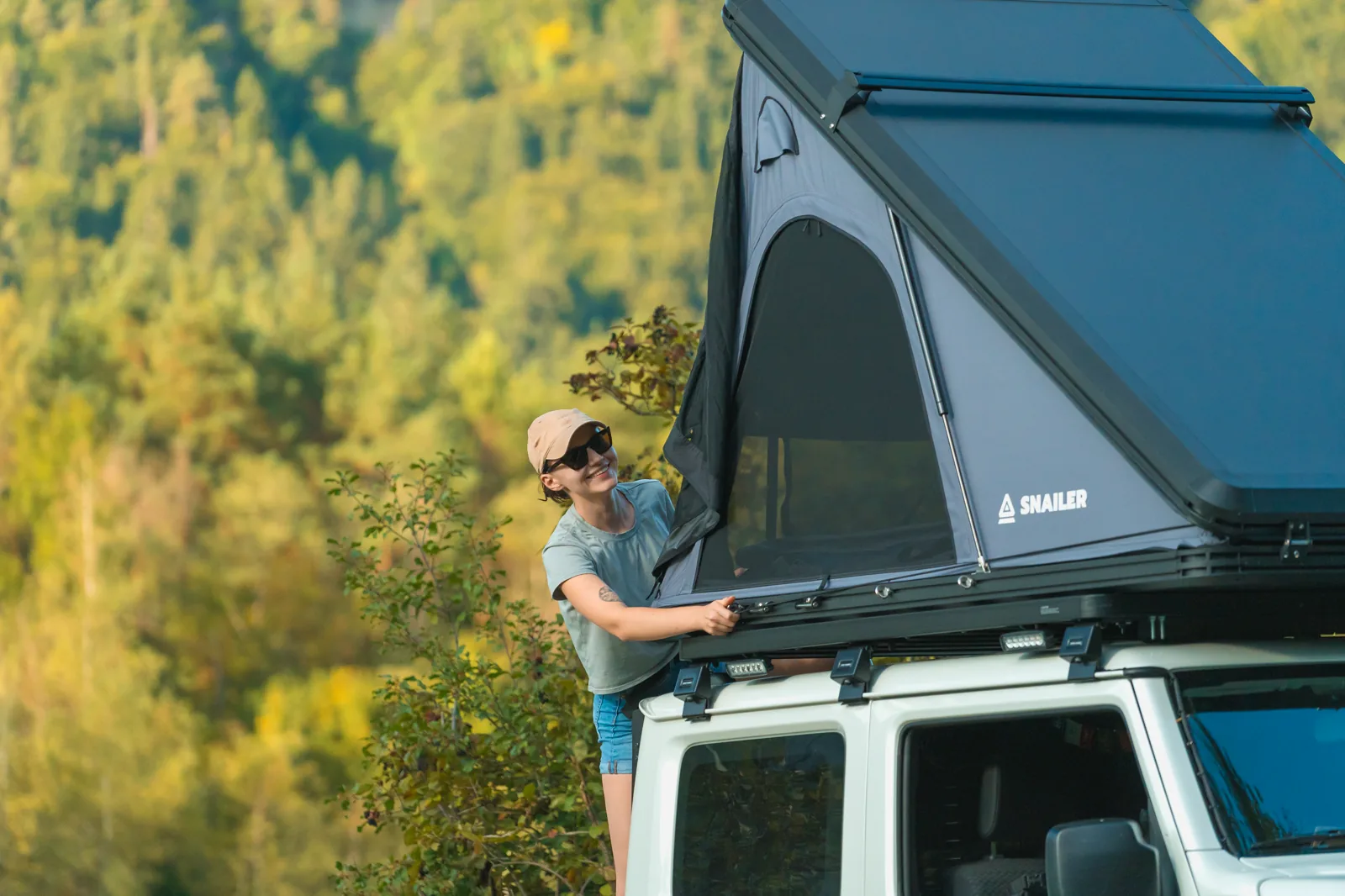 Woman opening a SNAILER hard-shell roof tent on an off-road vehicle in the mountains